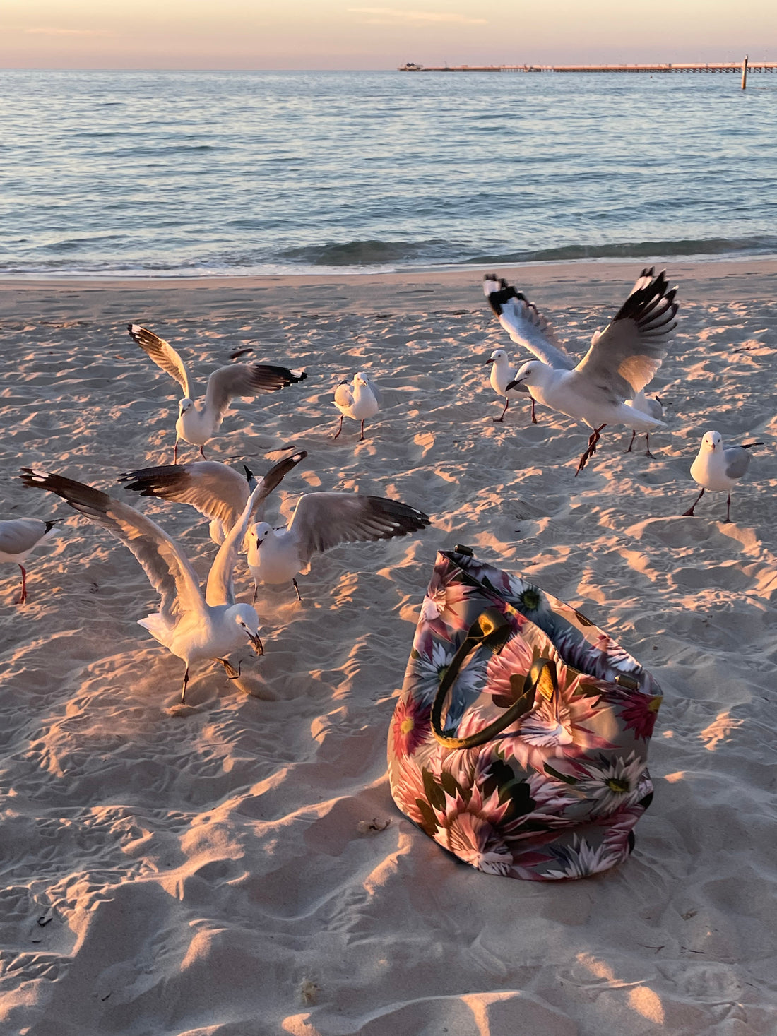 large water resistant, wildflower, canvas tote bag on the beach surrounded by flying seagulls. It is evening time and the sea is calm and the sand is full of footprints.