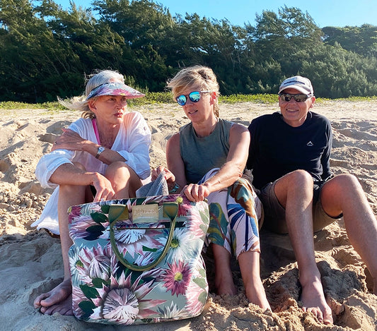 Three people sitting on a beach with a large canvas Wild Things Lifestyle beach bag in front of them. The bag has proteas on it and is called Bernadette
