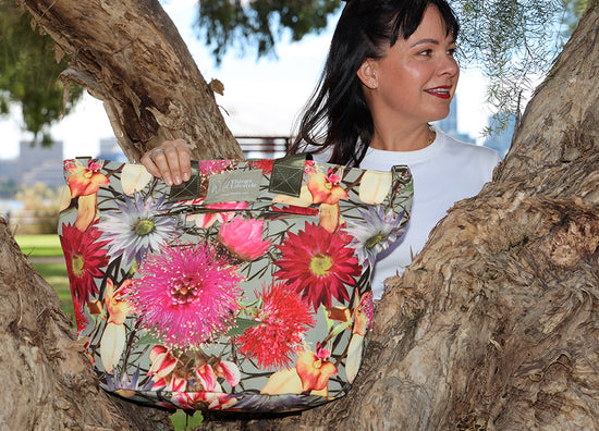 Lady standing in the fork of a tree in the park holding onto a Wild Things Lifestyle Tote Bag. She is looking to the side and smiling. This large ladies handbag is adorned with a medley of Australian wildflowers. The lady has dark hair.